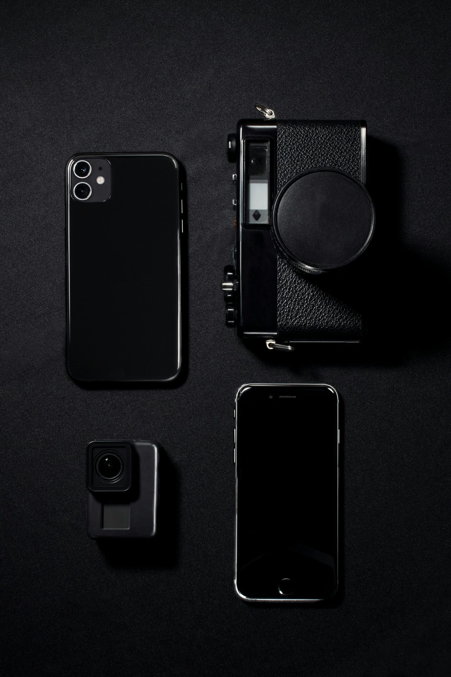 Minimalist flat lay of black gadgets including phones and a camera against a dark background.